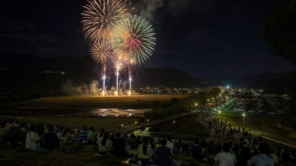 沼田市運動公園の来場者席から山あいの花火と帰路の歩行導線を望むAI生成の編集用画像
